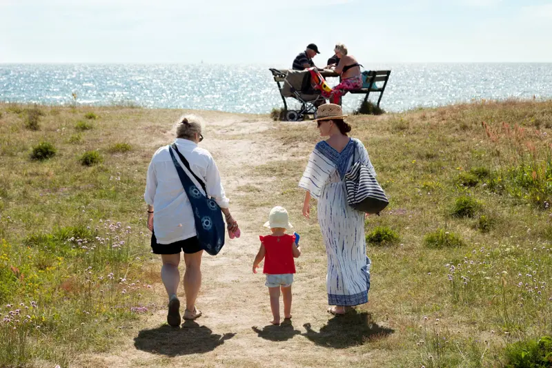 Two women and a toddler are walking towards the water front. They are in a meadow. In the distance, a couple of people are sitting on a bench with a table in front of it. It is summer. 