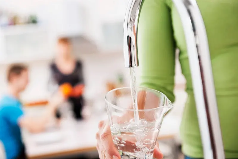 En hand håller i ett glas som fylls med vatten under en kran, personer sittandes vid ett bord i bakgrunden.