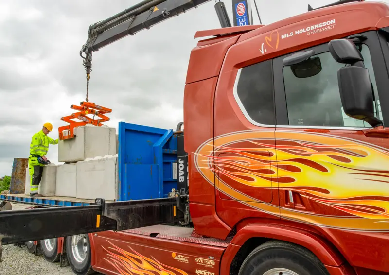 Person utför lastning av byggvaror på lastbilsflak med kran, lastbilen är målad i orangea eldflammor